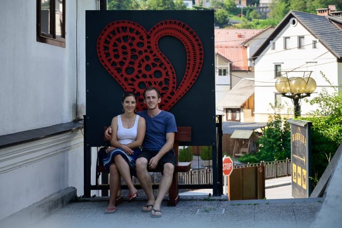 moški in ženska na klopci pred znakom za Idrijo - čipkasto srce