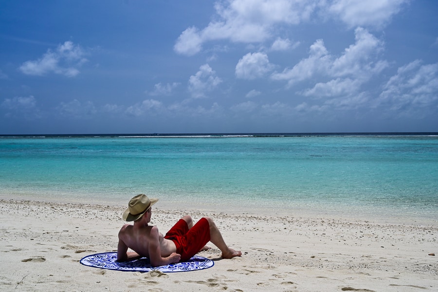 moški leži na okrogli brisači na peščeni plaži, turkizno morje v ozadju. Thoddoo, Maldivi