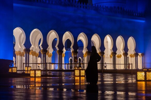 Nočna fotografija - mošeja Abu Dabi, ženska dolgi črni obleki hodi pred stebriščem