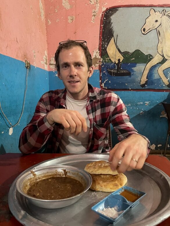 Moški pri mizi, pred seboj ima pladenj z rjavo juho (ful) ter kruhom