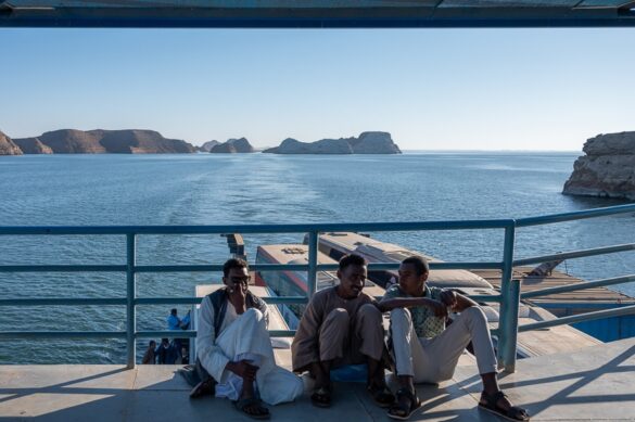 Trajekt čez jezero Nasser. Črni gospodje sedijo na ploščadi, spodaj so avtobusi, v ozadju je pogled na jezero Nasser